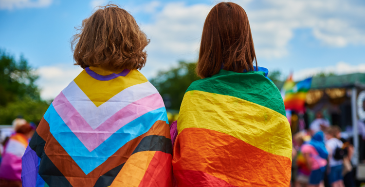 The backs of two people who are learning LGBTQIA+ flags around their shoulders.