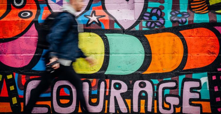 Young man walking past wall with graffiti saying Courage on it