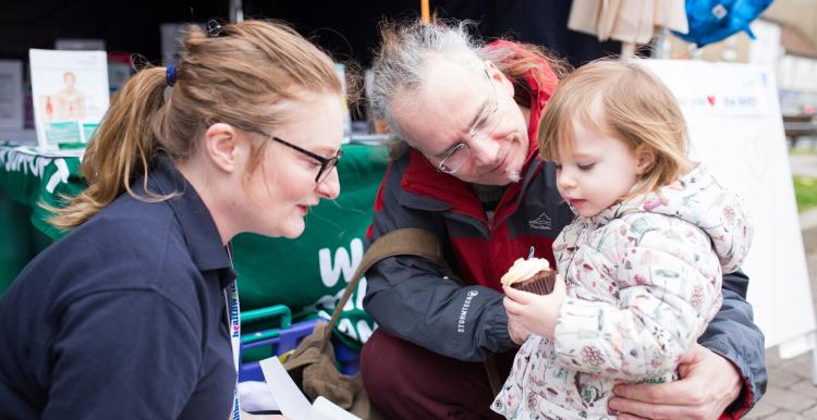 A man crouching down holding his daughter, who has a cupcake in her hand, with a Healthwatch volunteer also crouching down to speak to them 
