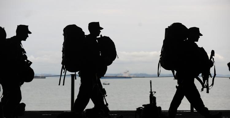 3 soldiers walking in a line with their backpack and weapons in front of a large body of water such as a lake