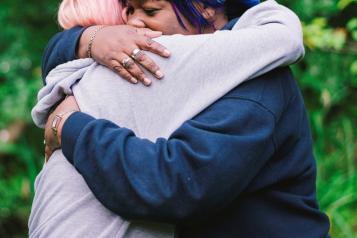 Two women hugging. One has pink hair and is wearing a grey jumper. The other is wearing a purple bandana and wearing a blue jumper. In the background, there are green leaves on a tree.