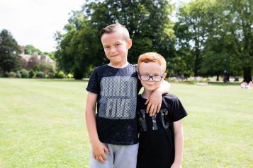 Two young boys, possibly brothers, standing together in a park outside in the summer