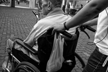 A black and white photo of an older man in a wheelchair being pushed by someone