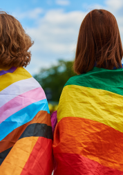 The backs of two people who are learning LGBTQIA+ flags around their shoulders.