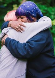 Two women hugging. One has pink hair and is wearing a grey jumper. The other is wearing a purple bandana and wearing a blue jumper. In the background, there are green leaves on a tree.