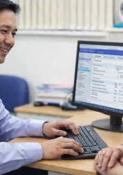 A healthcare professional sits at a desk smiling at an older woman while viewing patient information on a computer screen.