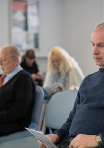 Photo of a middle aged man with people in the background all sitting in a waiting room in a medical setting
